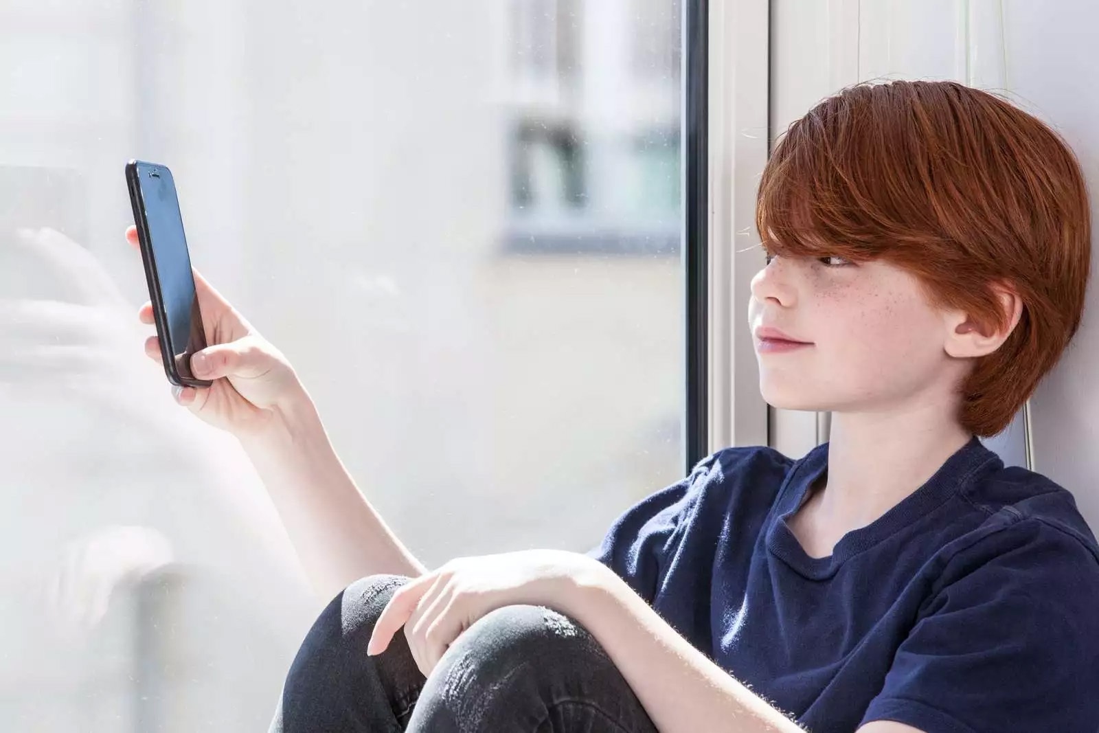 Rødhåret dreng sidder i vindueskam og kigger på en telefon.