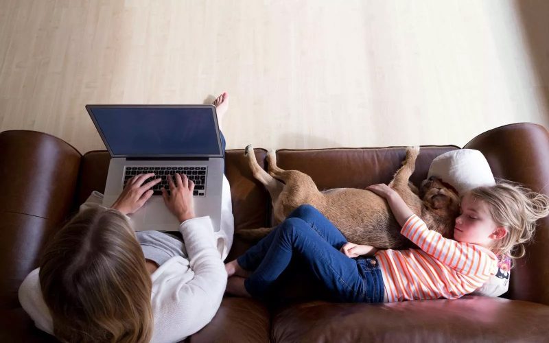 En kvinde sidder på en sofa med sin computer mens et barn sover ved siden af