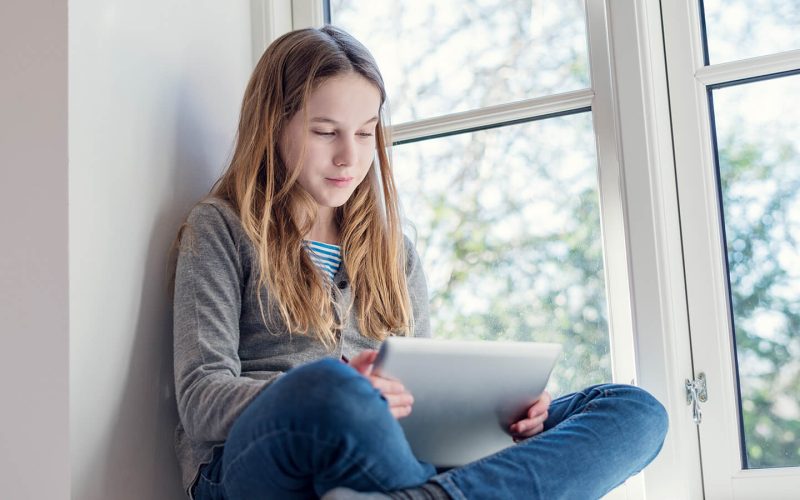 Pige sidder i vindueskarm med en tablet